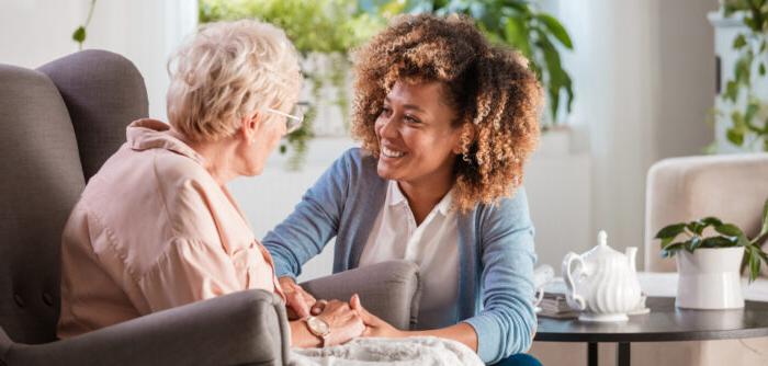 Female home caregiver talking with senior woman, sitting in living room and listening to her carefully.