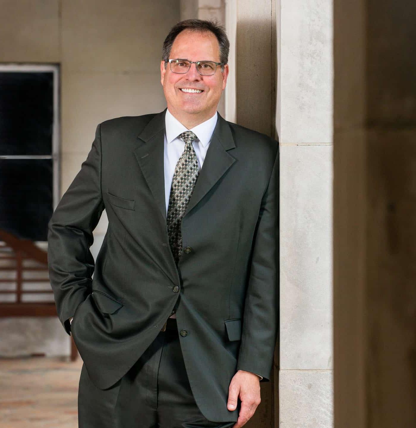 A smiling man in a dark suit leans casually against a stone pillar, posing in a formal yet relaxed manner.