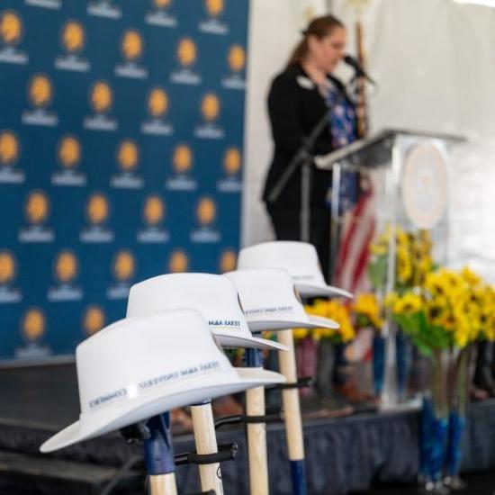 A woman speaks at a podium in the background, with an arrangement of sunflowers next to her. In the forefront, several ceremonial shovels with white hard hats labeled "Texas A&M University - Commerce" are arranged.