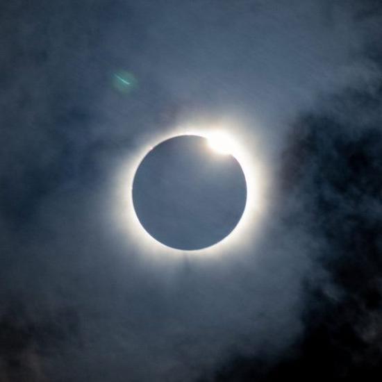 A solar eclipse is seen with the moon almost completely covering the sun. A bright ring of sunlight forms a halo around the black silhouette of the moon, with a small part of the sun peeking out from the top left. Dark clouds partially obscure the view.