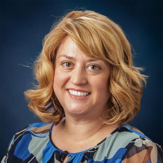 A smiling woman with shoulder length blonde hair, wearing a multicolor shirt, is set against a dark blue background.