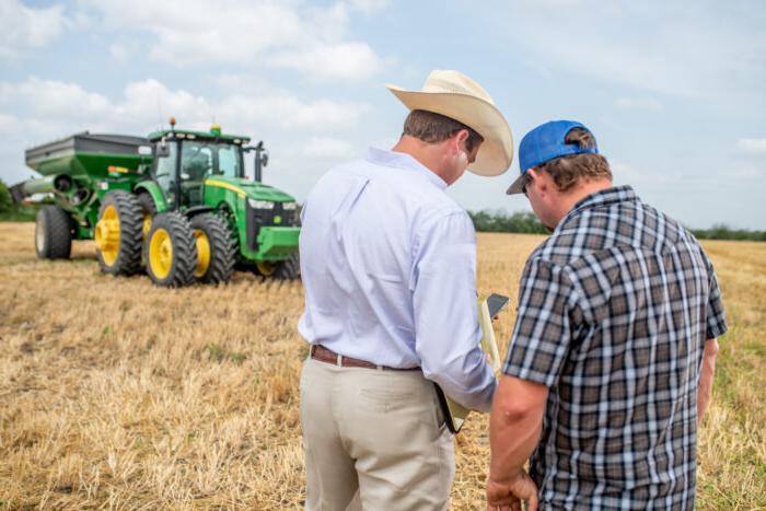 Two men, one in a cowboy hat and another in a blue cap, are standing in a field beside a large green tractor. They appear to be discussing something while looking at a cell phone.