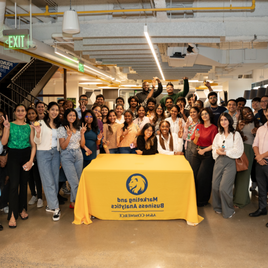 A large group of people pose for a photo around a table covered with a cloth featuring the logo for the department of marketing and business analytics at A&M-Commerce.